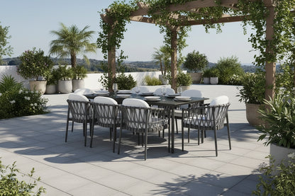 Outdoor dining set with a rectangular dark table and six woven gray chairs with white cushions, placed on a light stone patio beside a swimming pool with palm trees and a distant landscape.