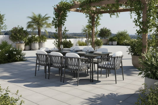 Outdoor dining set with a rectangular dark table and six woven gray chairs with white cushions, placed on a light stone patio beside a swimming pool with palm trees and a distant landscape.
